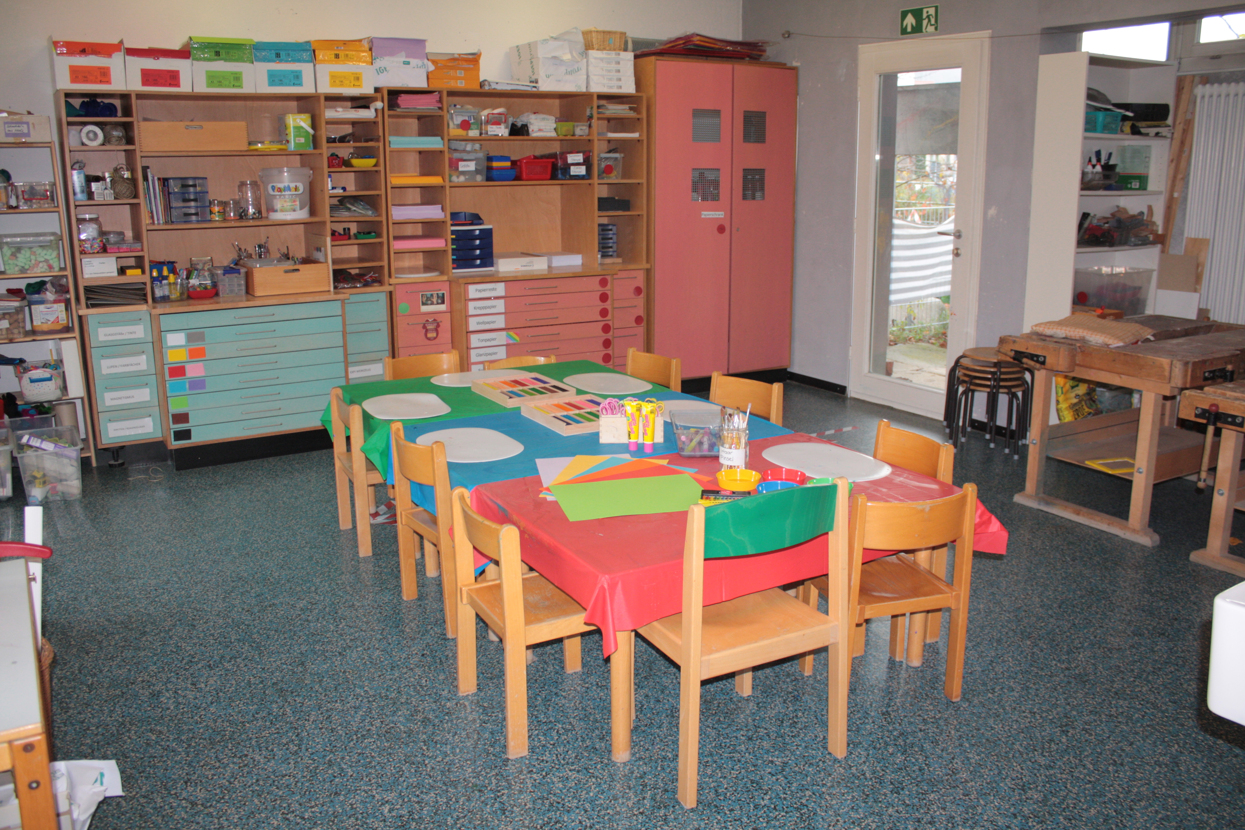 Rechts stehen zwei Werkbänke aus Holz, an der hinteren Wand ist ein großes Regal mit verschiedenen Fächern und Schubladen mit Bastelmaterial und Farben erkennbar. In der Mitte des Raumes steht ein rechteckiger Tisch mit Bastelmaterial.