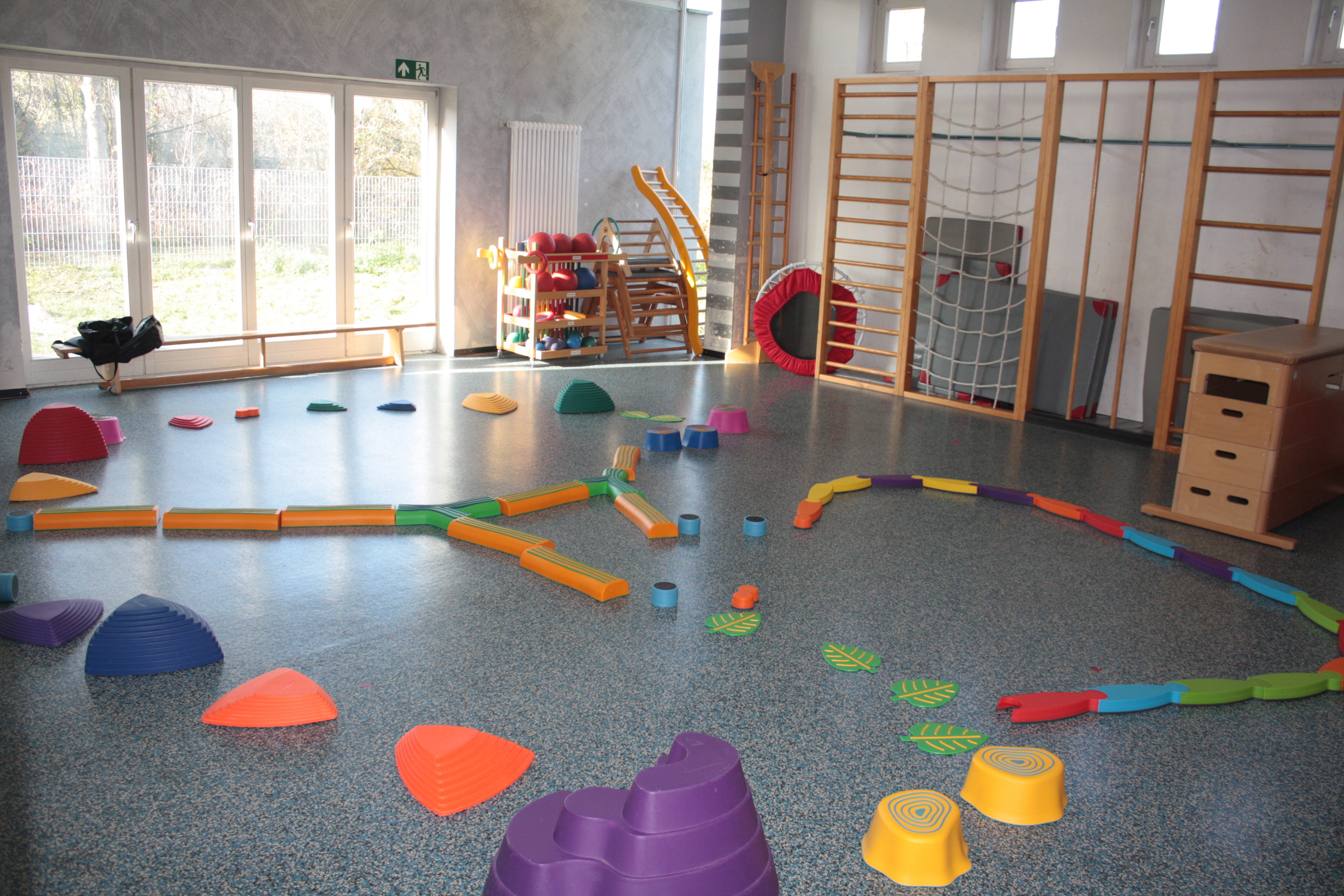 Das Bild zeigt die Turnhalle mit einer Balancierlandschaft. Im Hintergrund ist eine Sprossen- und Kletterwand, Bälle und ein Trampolin abgebildet.