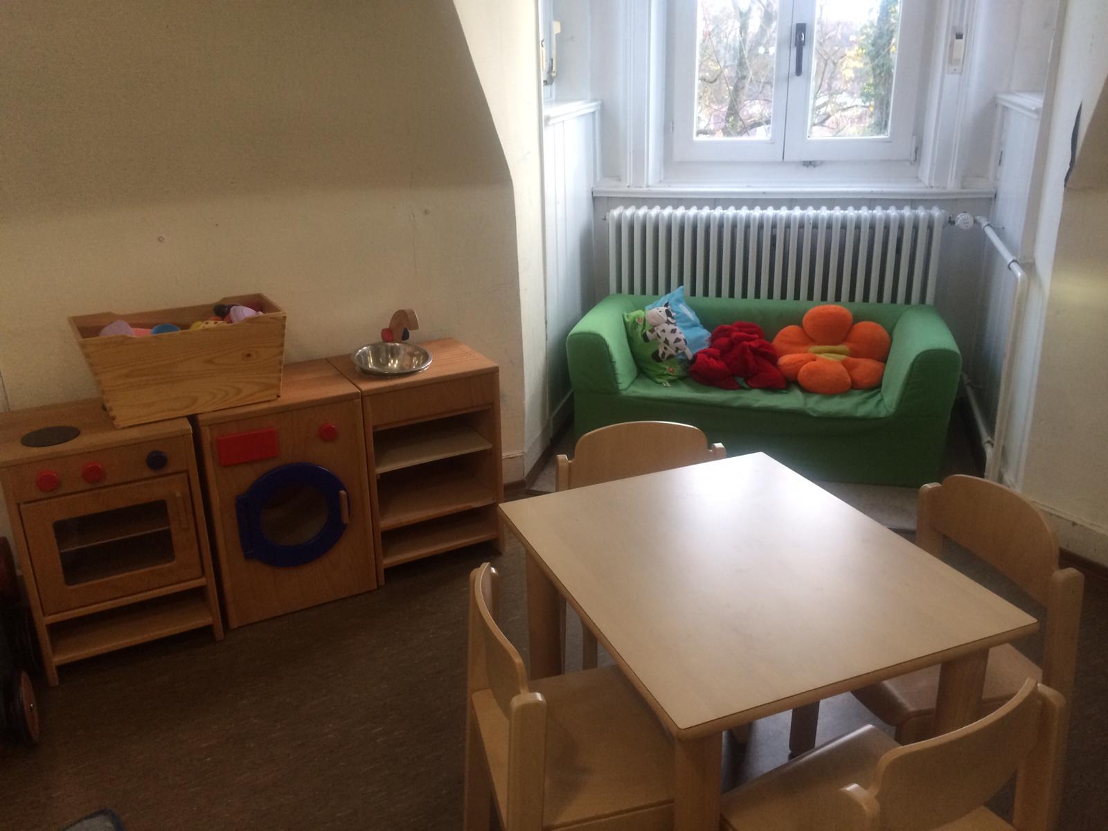 Das Bild zeigt die Puppenecke des Kinderhauses. Es ist eine Holzkinderküche, eine Holzwaschmaschine, ein kleines Sofa am Fenster zu sehen. Im Vordergrund ist ein kleiner Tisch erkennbar.