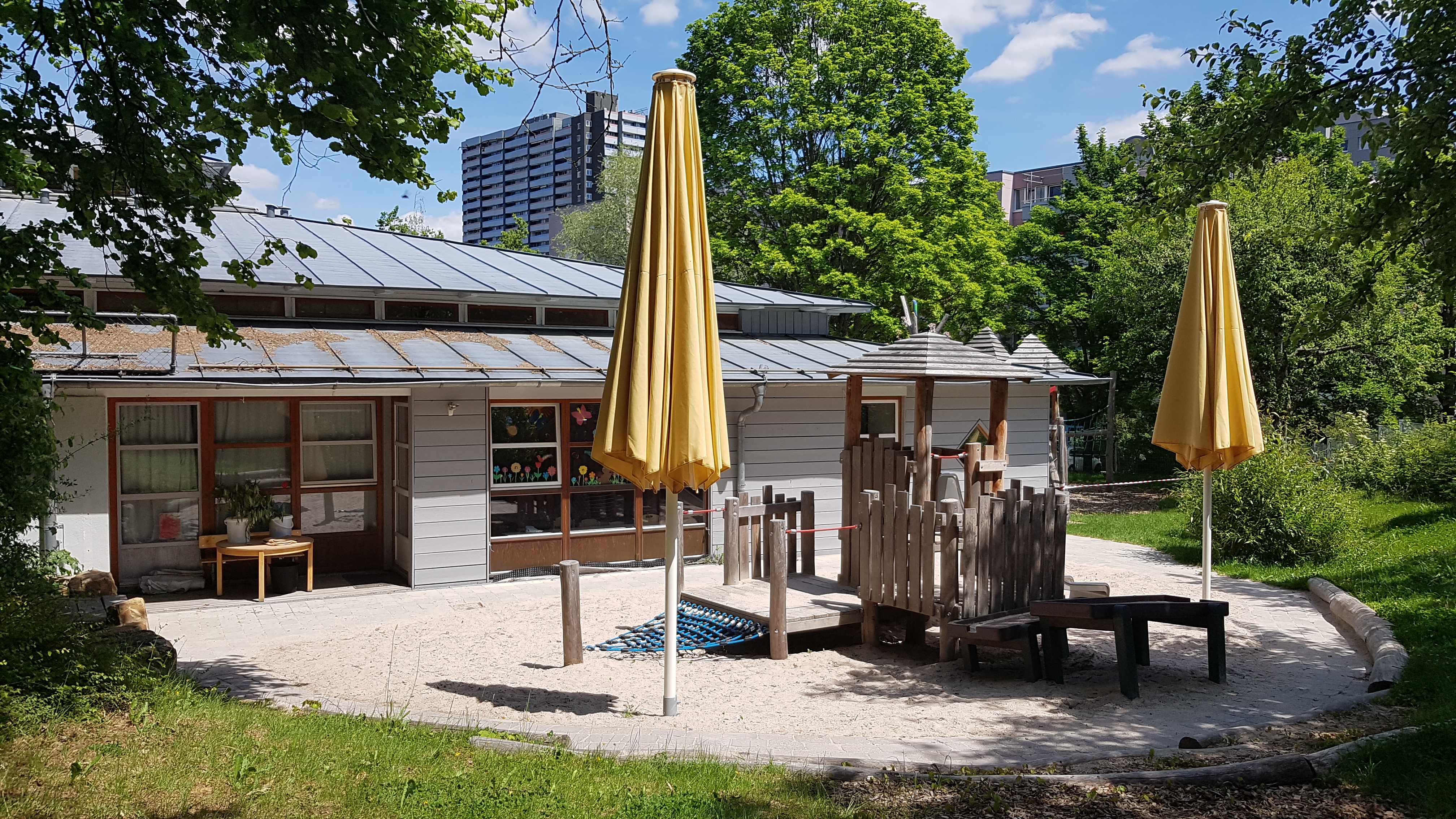 Das Bild zeigt einen weiteren Teil des Gartens. Ein großer Sandkasten mit kleinem Spielhaus aus Holz mit Matschtisch und kleiner Rutsche. Zwei große gelbe Sonnenschirme spenden Schatten.