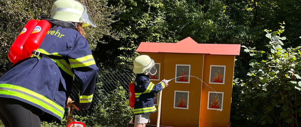 Ein Kind und ein Erwachsener löschen mit einem Feuerwehrschlauch ein künstliches Feuer.