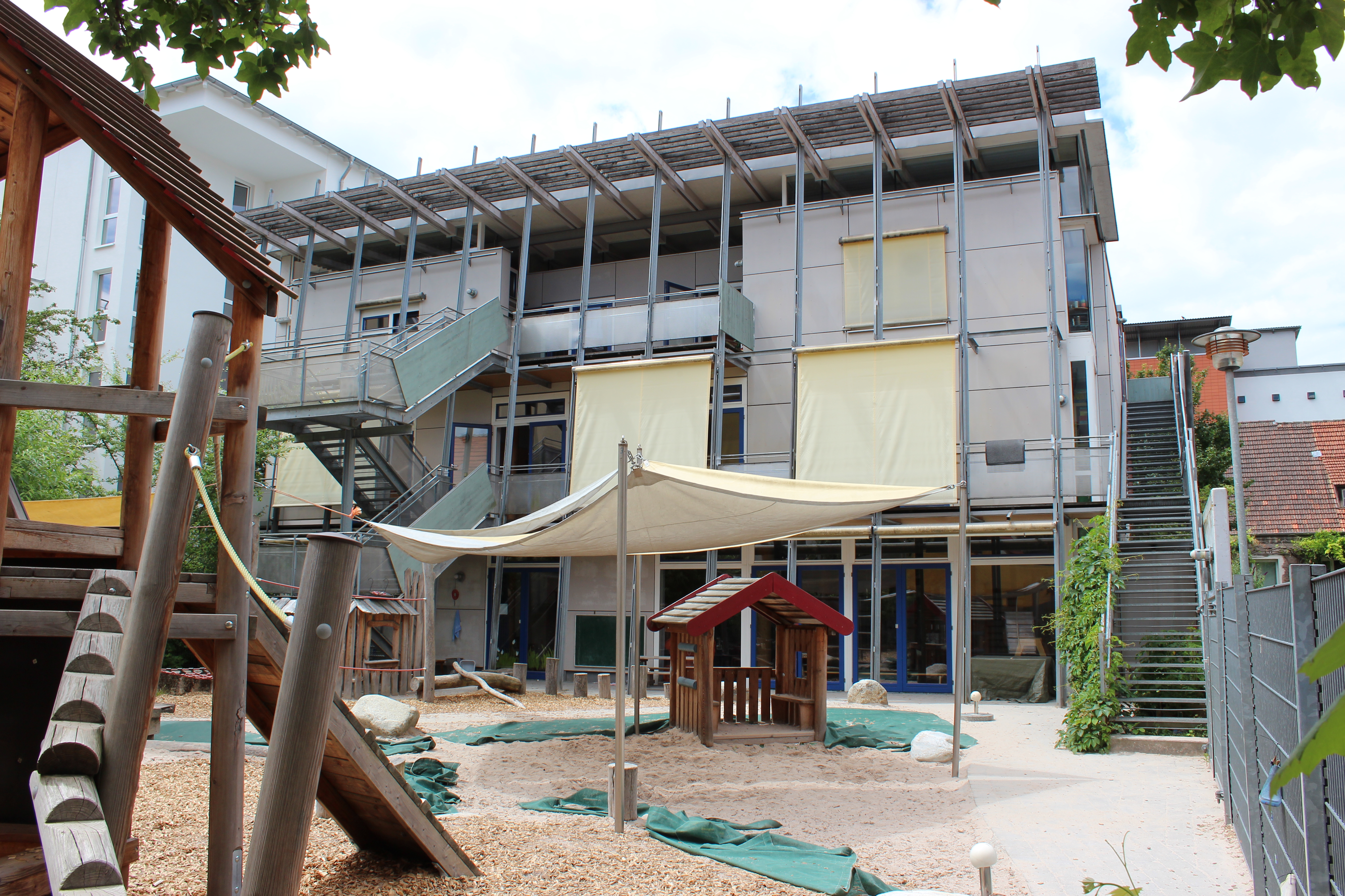 Das Bild zeigt den Garten des Kinderhaus. Es sind ein Spiel- und Kletterhaus, ein Sandkasten mit Sonnensegel und ein kleines Spielhaus aus Holz zu sehen.