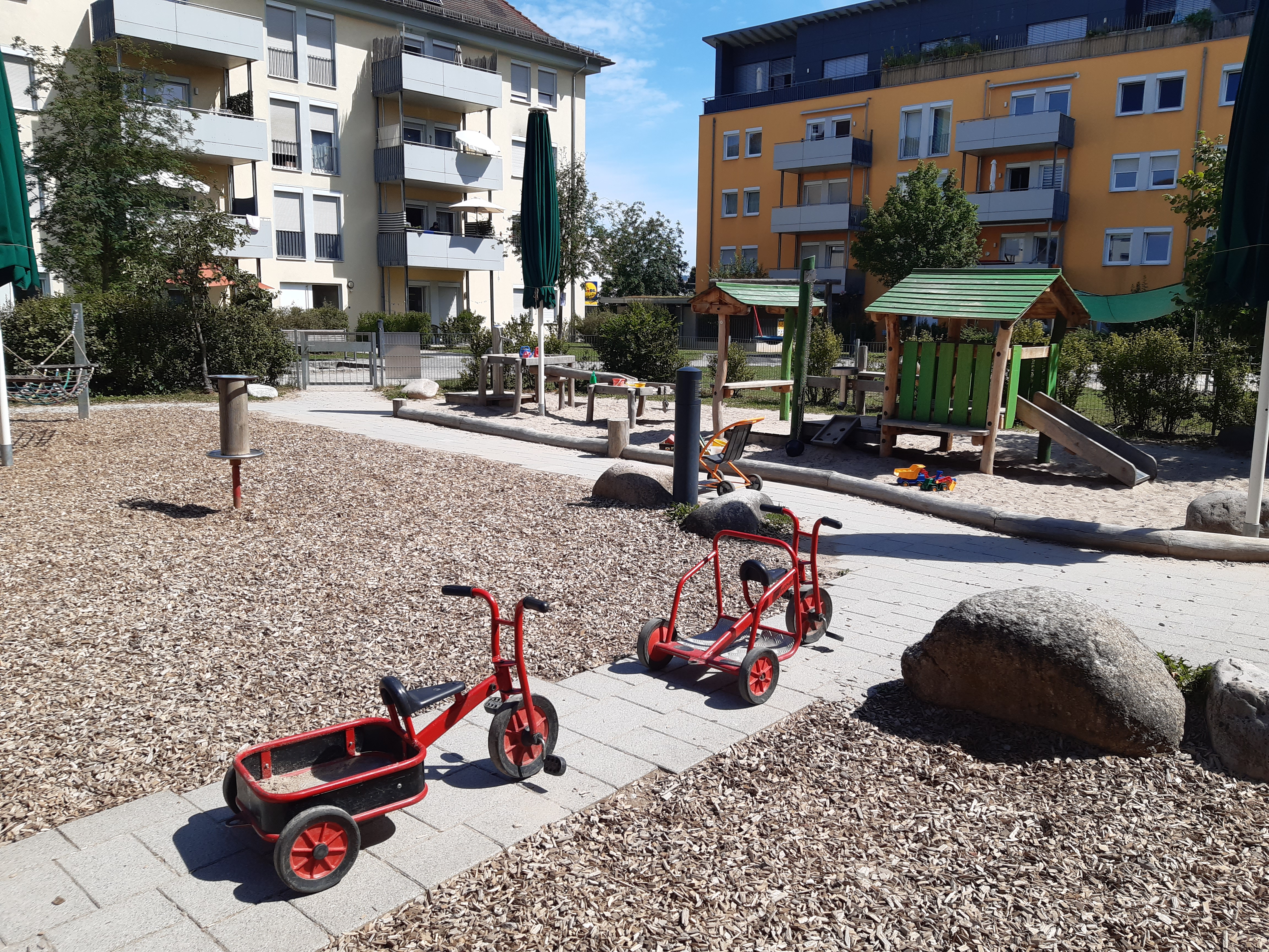 Auf dem Bild ist ein großer Sandkasten mit Spiel Häusern zu sehen. Im Vordergrund sind zwei rote Dreiräder abgebildet.