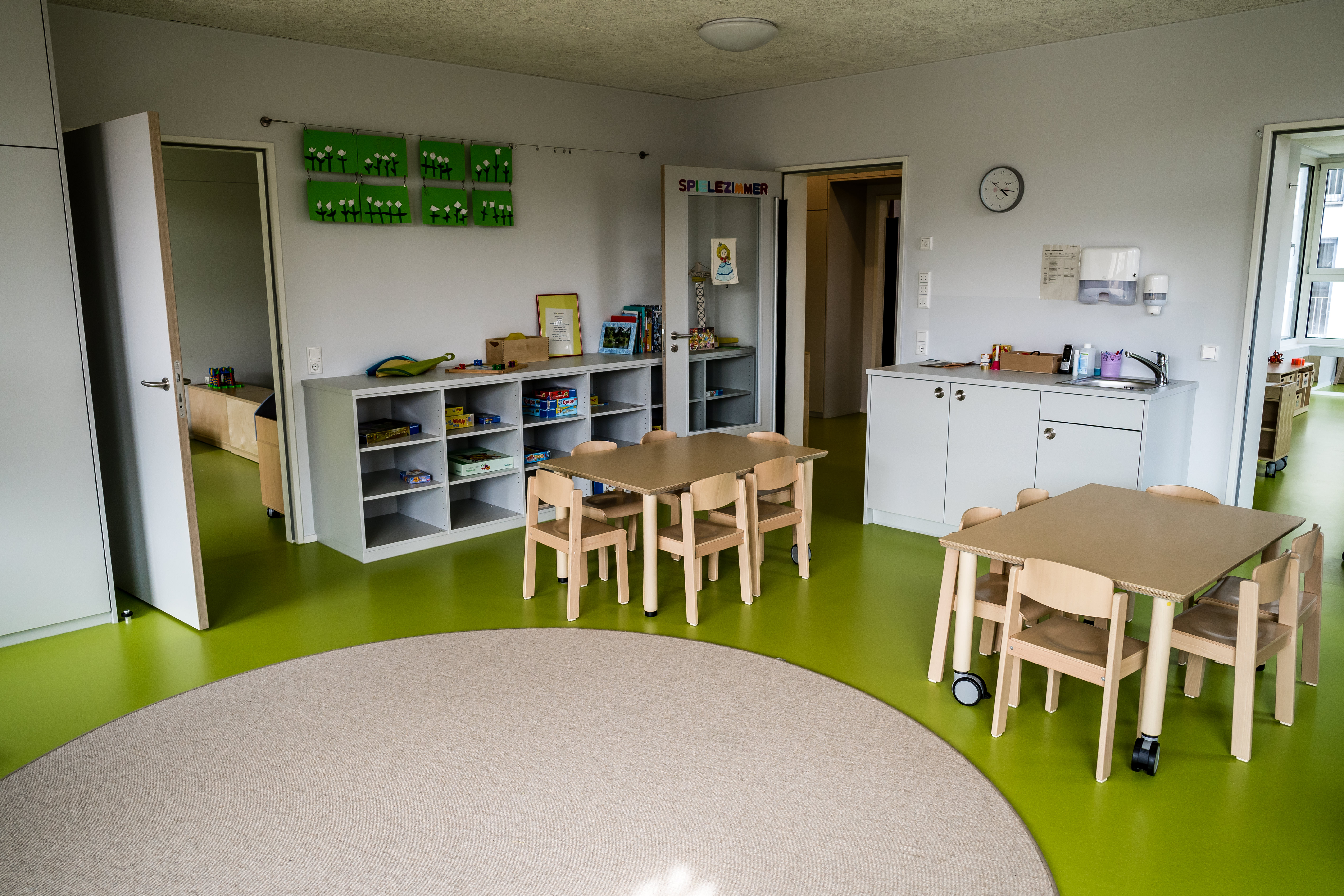 Das Bild zeigt den Gruppenraum im Kindergarten. Es ist links ein Regal mit Spielsachen und davor zwei Tische mit Stühlen zu sehen.