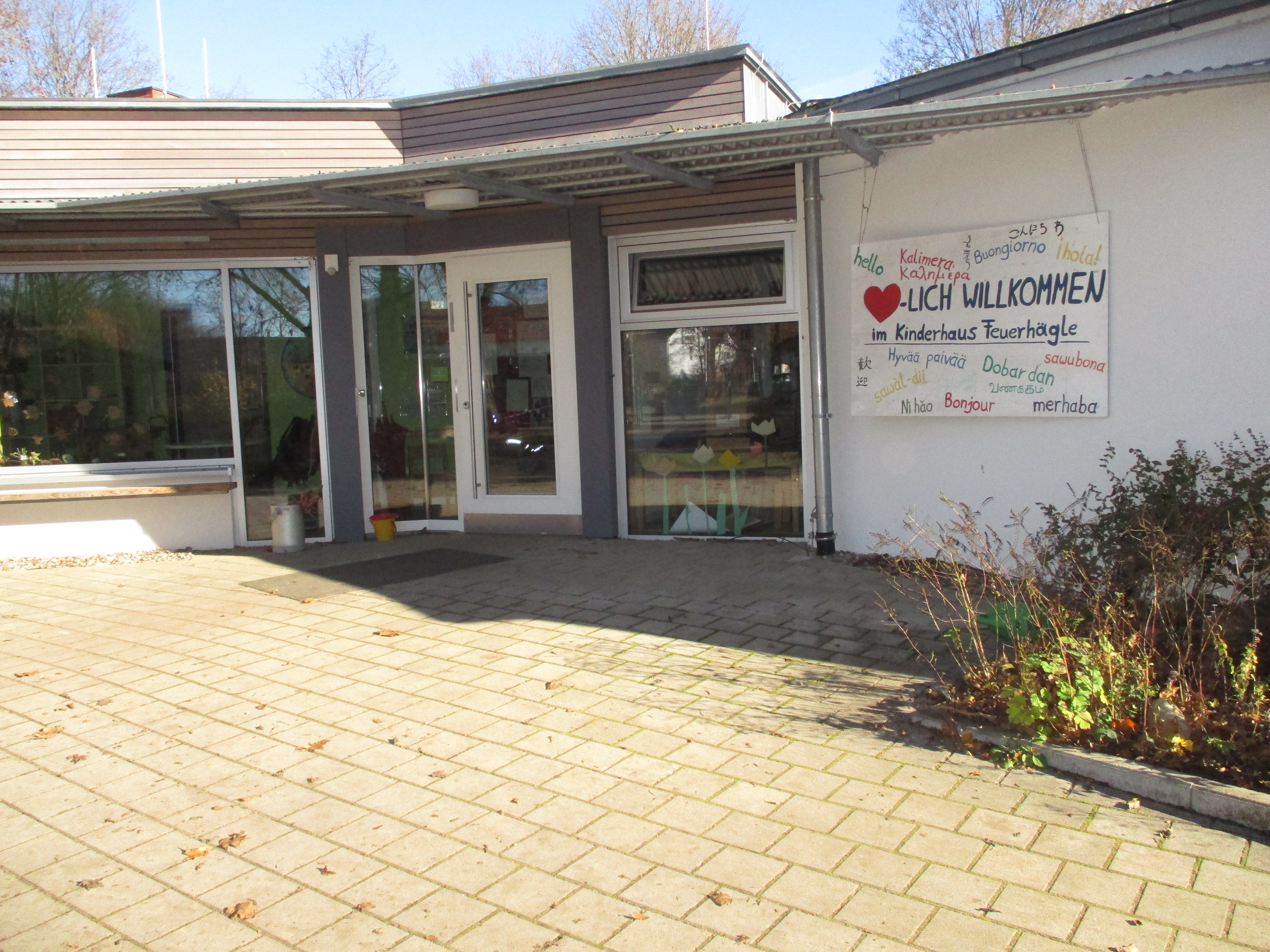 Das Bild zeigt den Eingangsbereich des Kinderhaus Feuerhägle. Rechts neben der Tür hängt ein Willkommensplakat. Es sind verschiedene Sprachen darauf abgebildet.