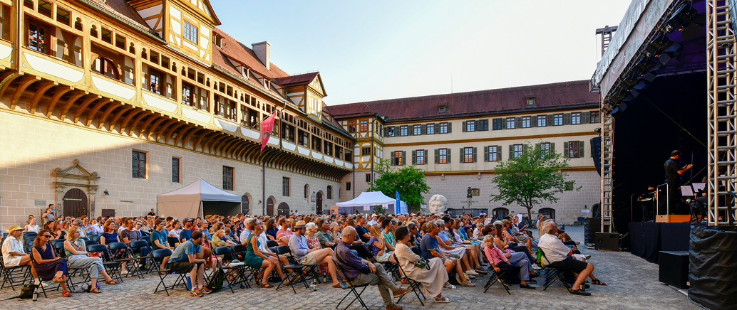 Das Publikum eines Schlosshofkonzertes vom letzten Jahr 2022 sitzt zur Bühne gerichtet auf dem Schlosshof und wurde von der Seite fotografiert.