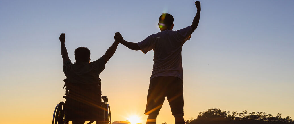 Ein Mann im Rollstuhl neben einem stehenden Mann vor einem Sonnenuntergang