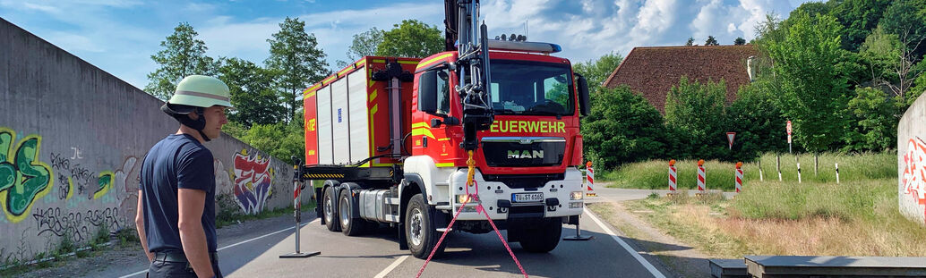 Feuwehrmann im Einsatz auf einer Straße. Im Hintergrund steht ein Feuerwehrauto.