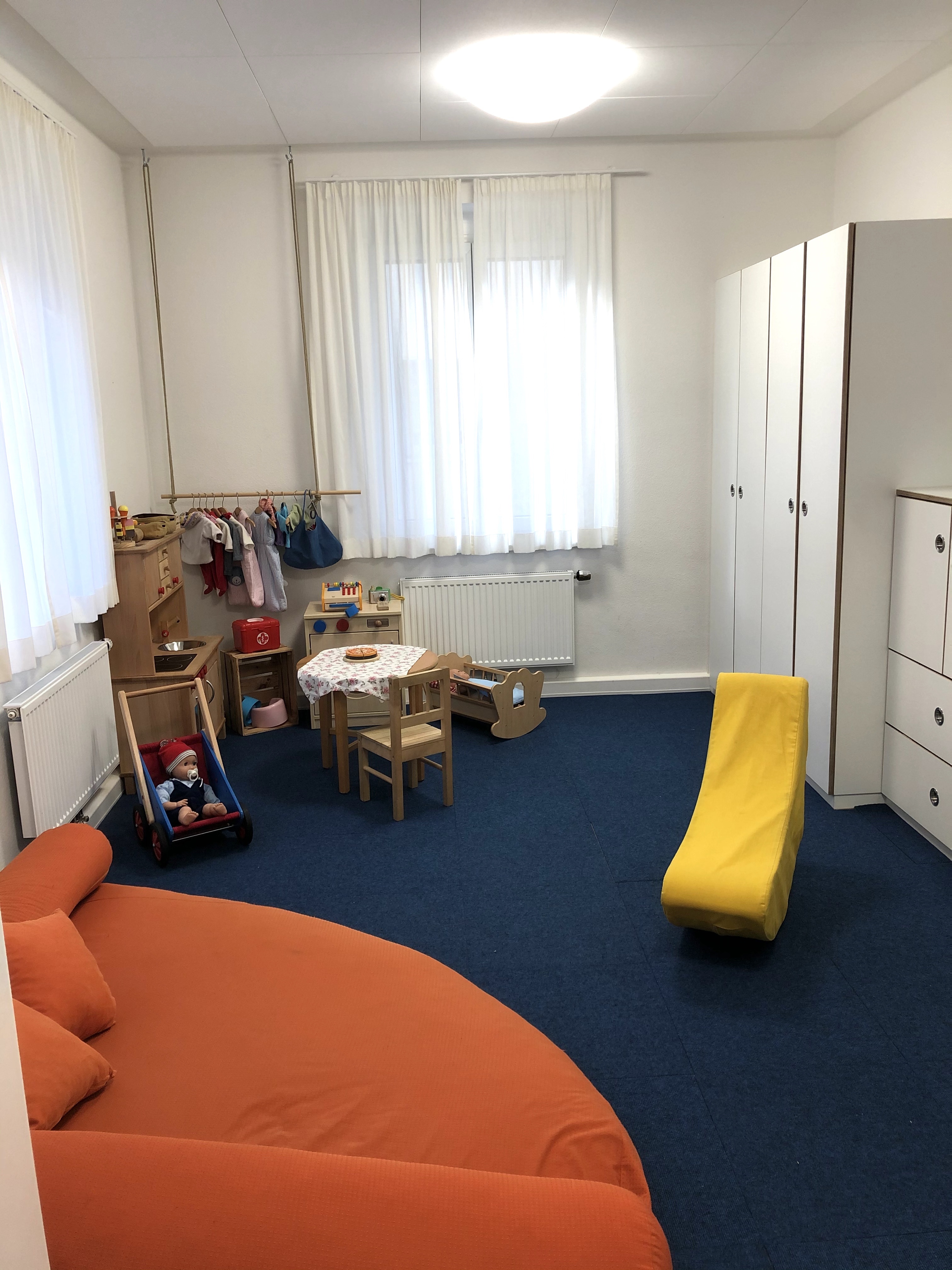 An der rechten Wand sind weiße Schränke eingebaut. In der hinteren linken Ecke steht eine Kinderküche, sowie eine  Kleiderstange für Puppenkleider. davor steht ein kleiner Tisch, eine Puppenwiege und ein Puppenkinderwagen. Vorne links ist ein Sofa.