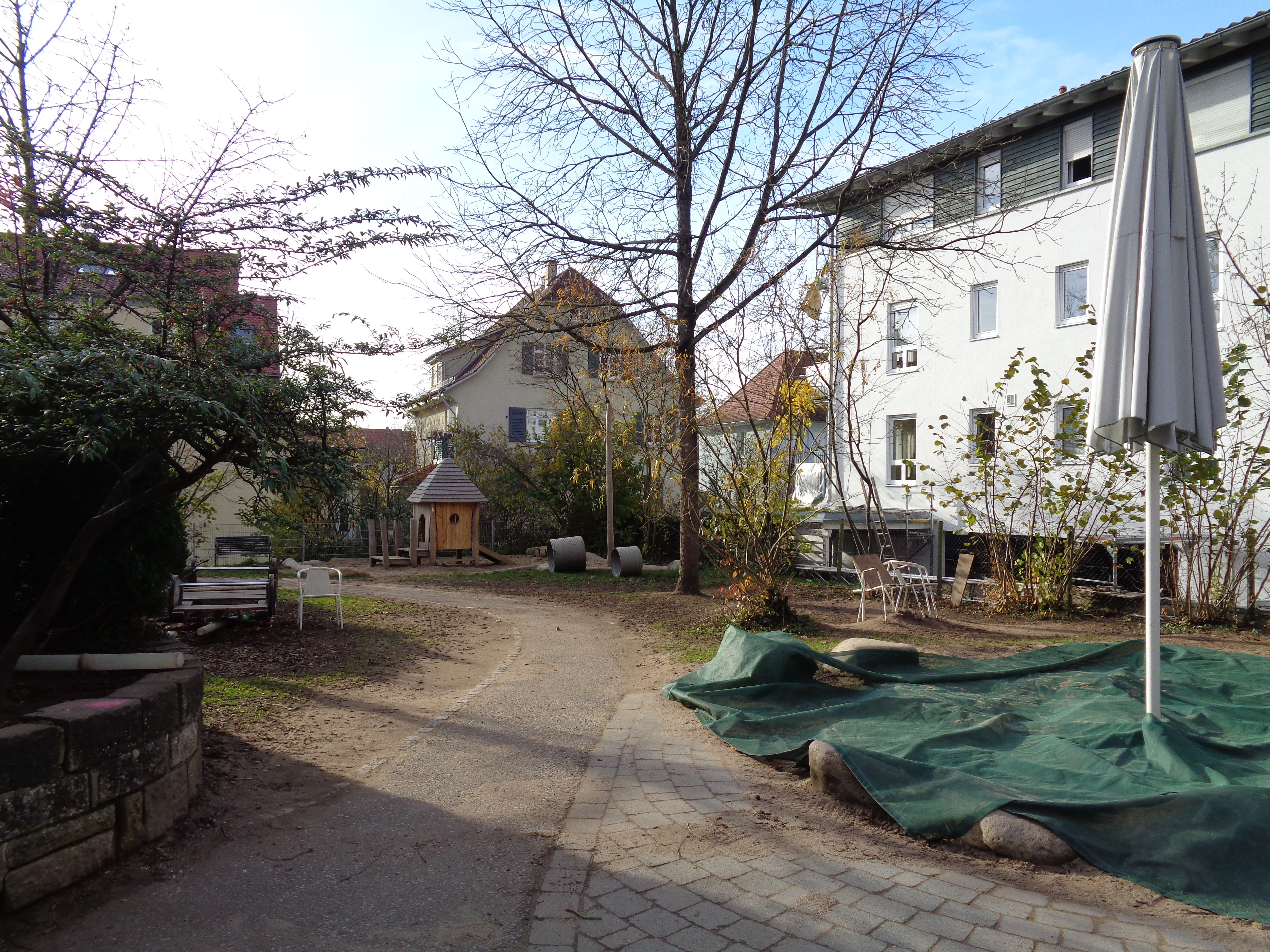 Das Bild zeigt rechts einen abgedeckten Sandkasten mit Sonnenschirm. im Hintergrund ist ein Spielhaus aus Holz erkennbar.