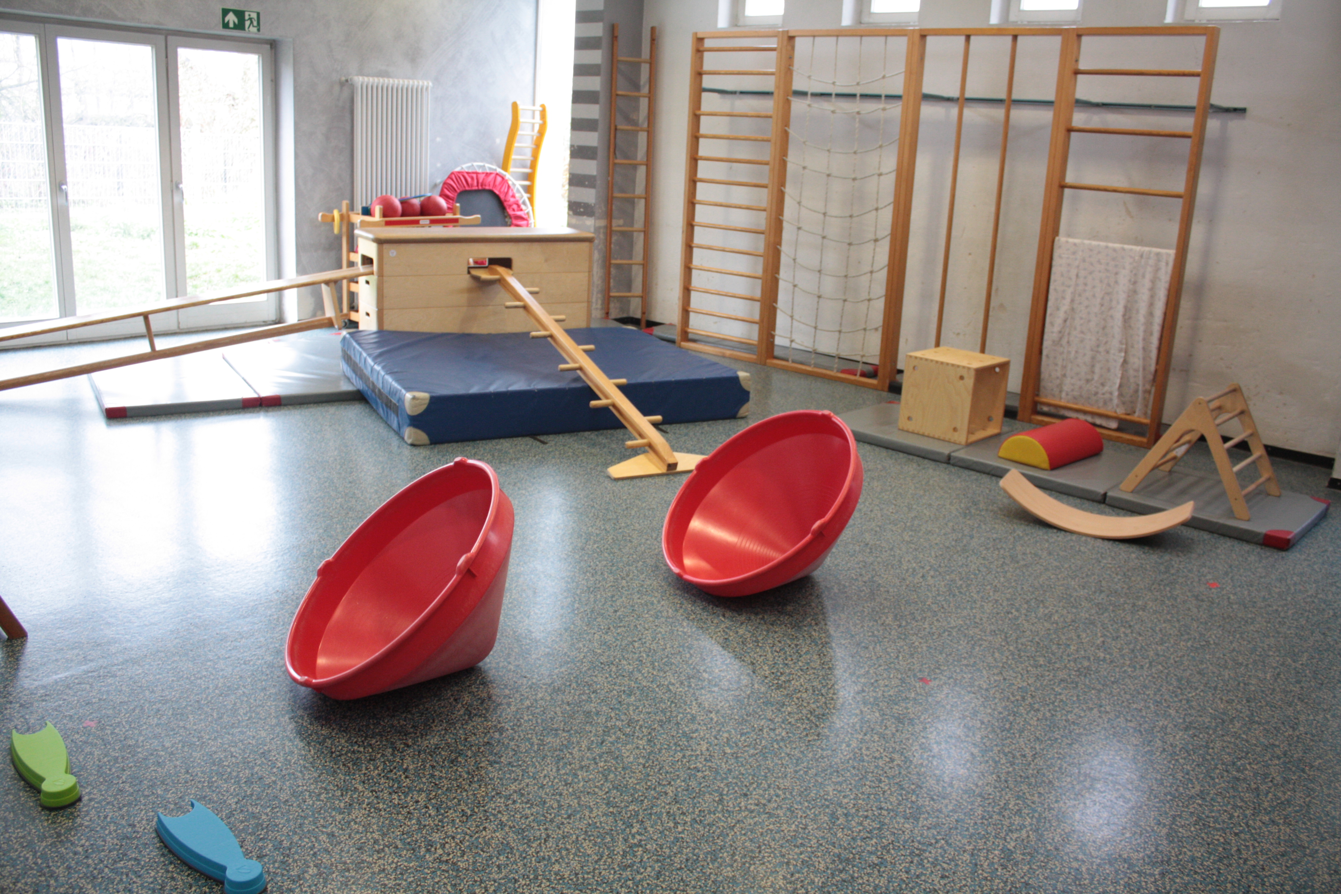 In der Turnhalle ist eine Bewegungslandschaft zum Klettern und Balancieren über Kästen und Bänke aufgebaut. Im Vordergrund sind zwei rote Kreisel erkennbar.