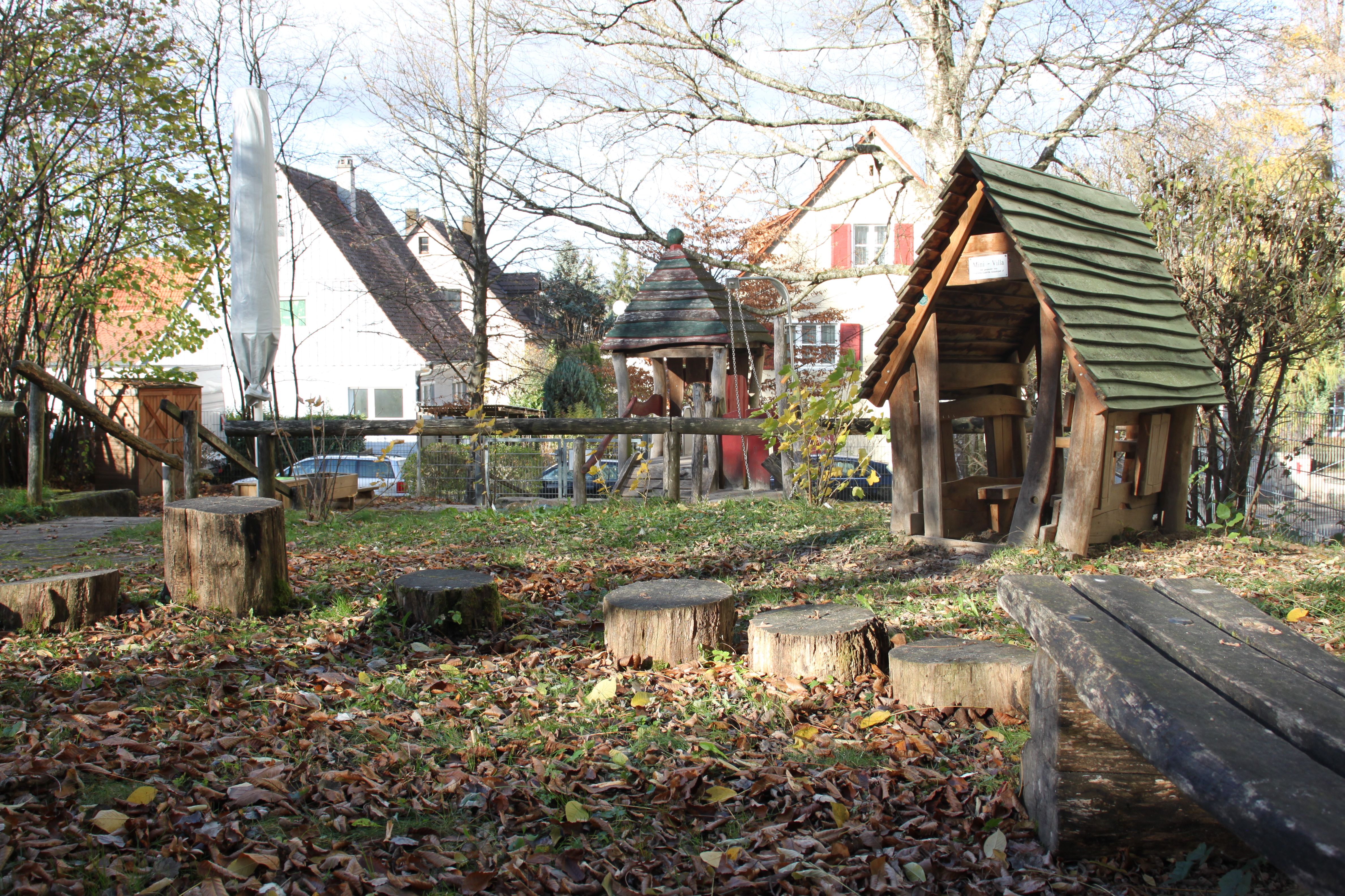 Das Bild zeit einen Ausschnitt des Gartens. Im Vordergrund dicke Holzstämme zum hüpfen. Dahinter ist ein Spielhaus aus Holz.