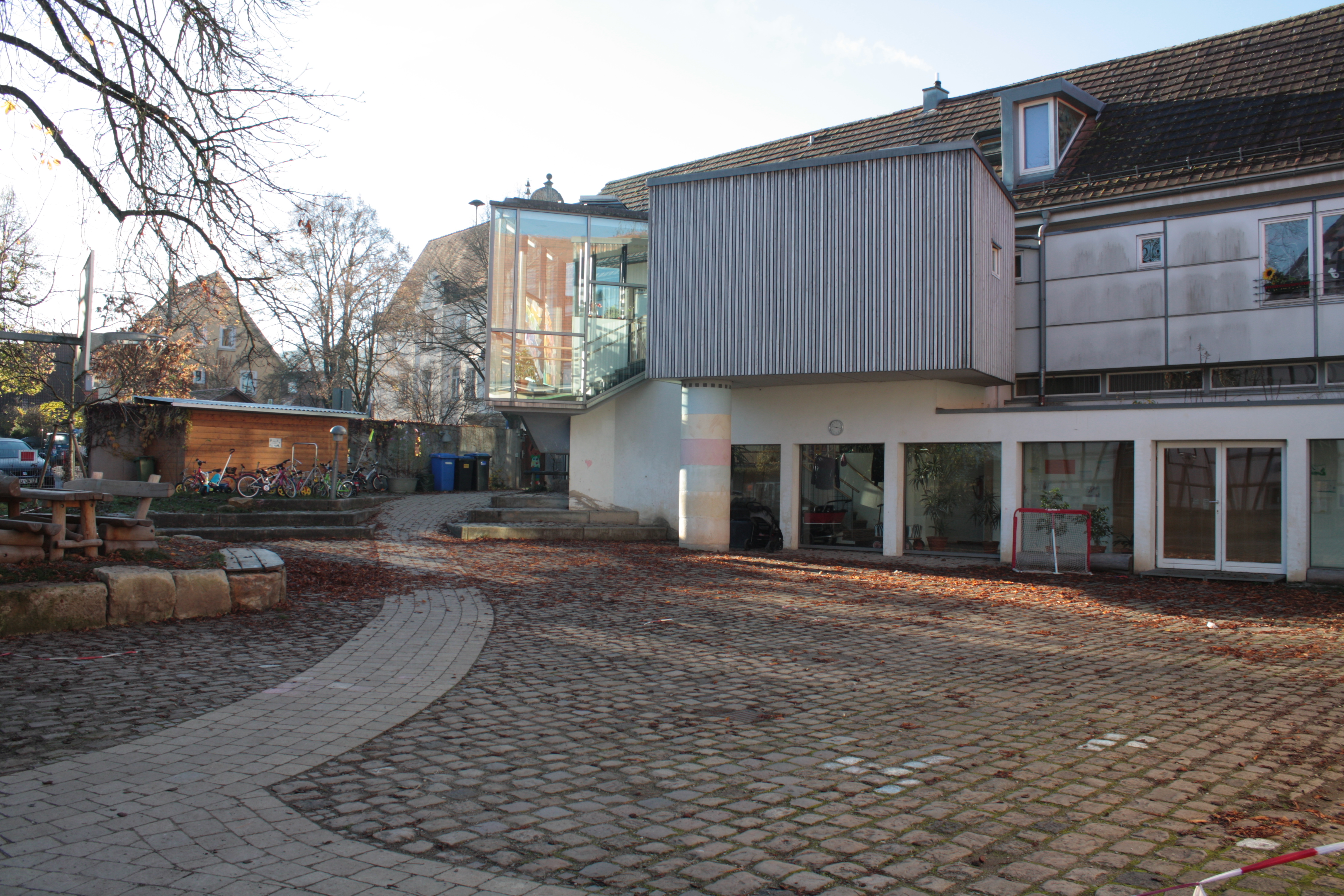 Auf dem Bild ist die Außenansicht des Kinderhauses Alte Mühle und im Vordergrund der gepflasterte Hof. Links ist eine Holzbank mit Tisch erkennbar.