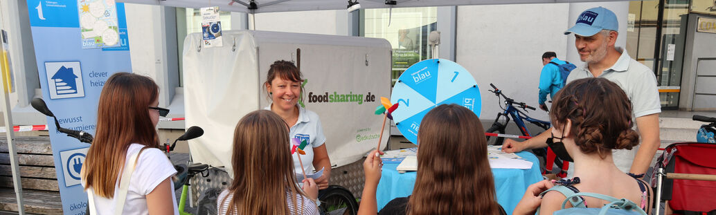 Ein Mann und eine Frau stehen an einem Infostand und erklären vier Mädchen etwas.