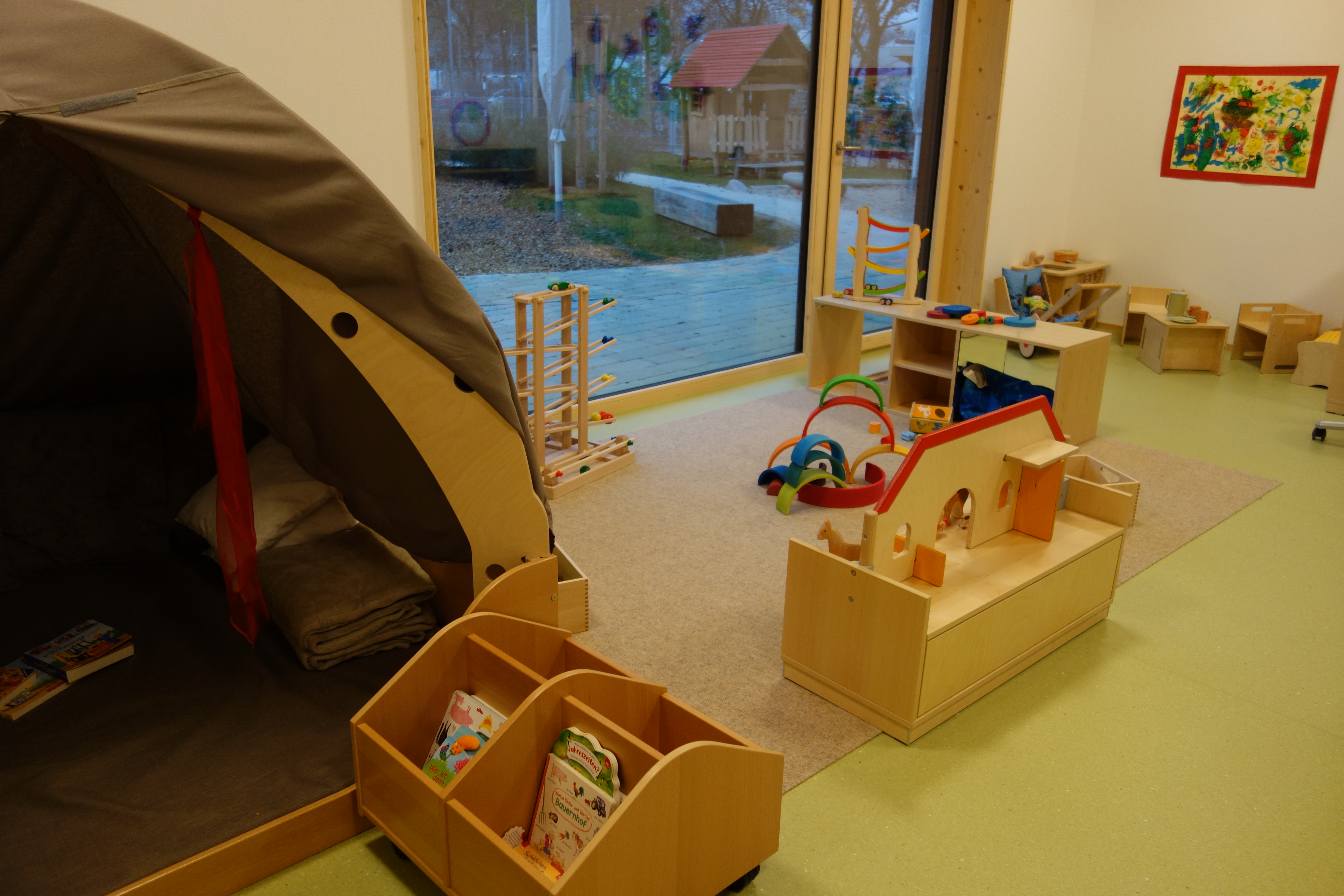 Das Bild zeigt den Gruppenraum der Krippenkinder. Links eine Kuschelhöhle, mit Bücherkisten davor. Im Hintergrund ist eine Bauecke und eine Rollenspielecke abgebildet.