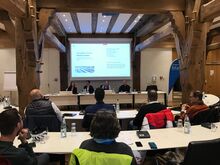 Menschen sitzen im Ratsaal im Rathaus und schauen auf eine Beamerleinwand