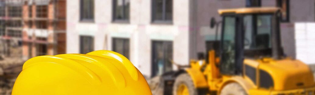 Im Vordergrund ein Bauhelm, im Hintergrund Bagger und Neubau