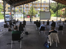 Menschen sitzen in einer Halle auf St&uuml;hlen, eine Frau steht vor ihnen und spricht