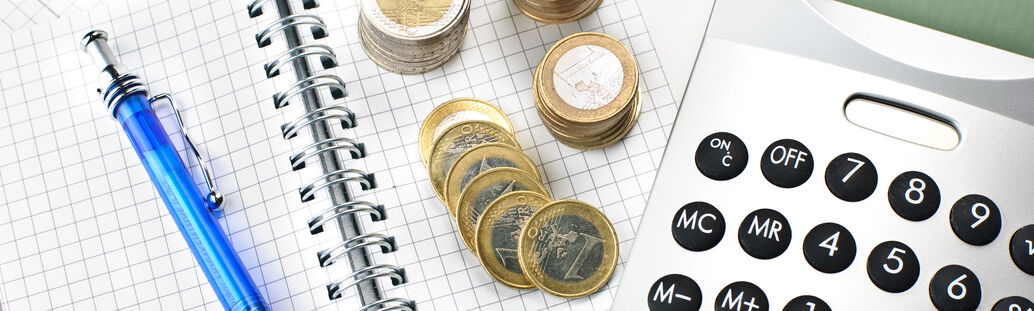 Auf einem karierten Notizblock liegen Eurom&uuml;nzen, ein Kugelschreiber und ein Taschenrechner.