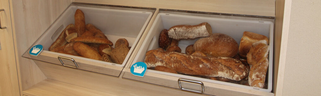 Brote, Baguettes und Br&ouml;tchen liegen in zwei gro&szlig;en Ablagen unter einer Schutzscheibe (Fairteiler).