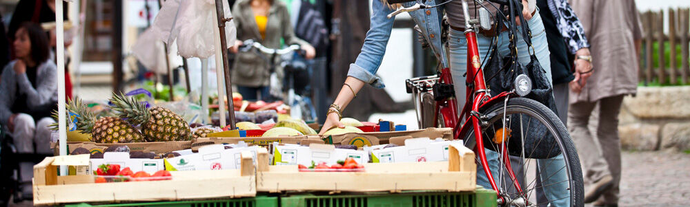 Eine Frau steht mir ihrem Fahrrad an einem Wochenmarktstand und greift nach Obst und Gem&uuml;se.